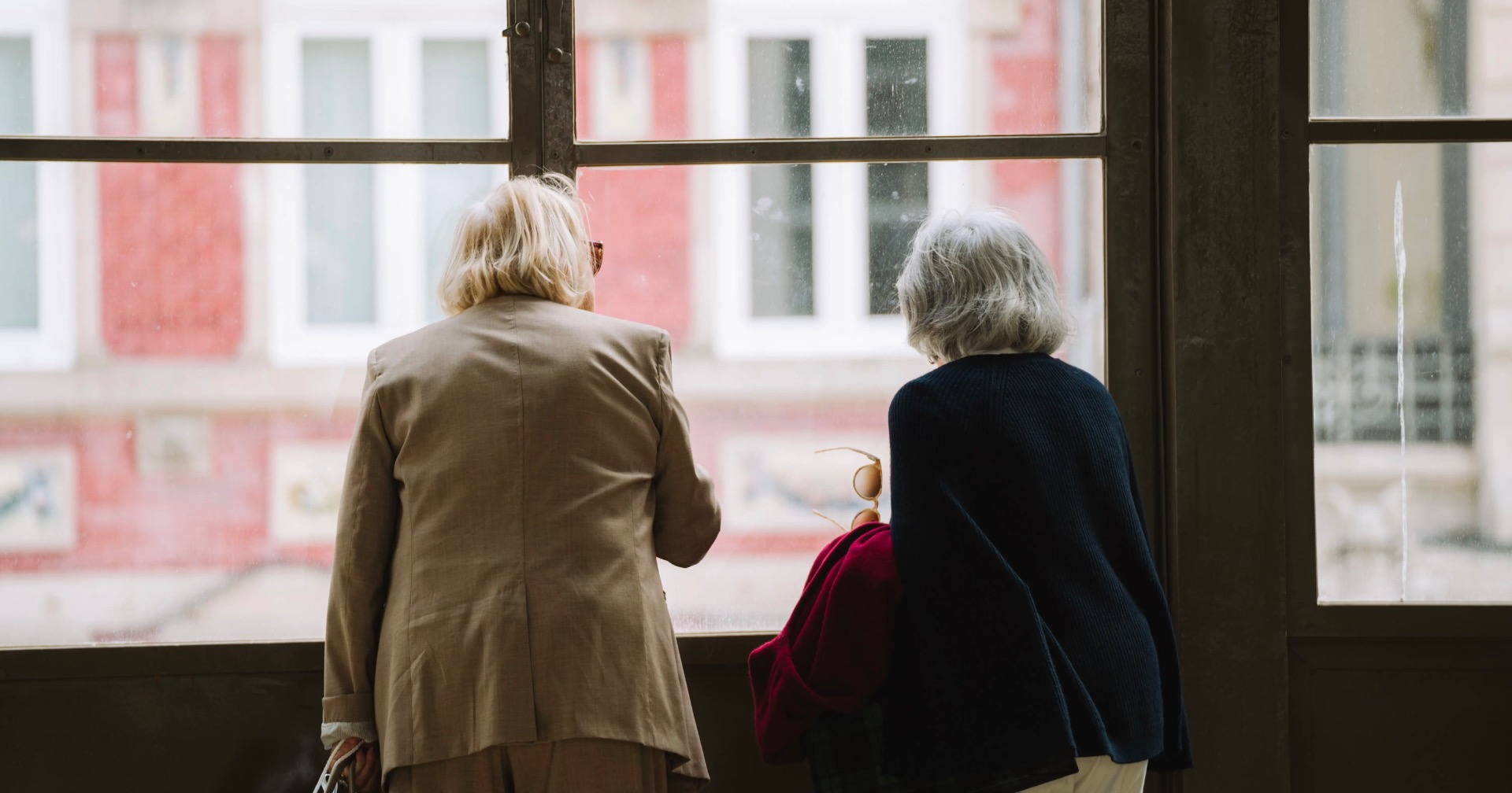 Dos mujeres mayores, de espaldas, observan a través de una ventana la fachada de un edificio de viviendas.