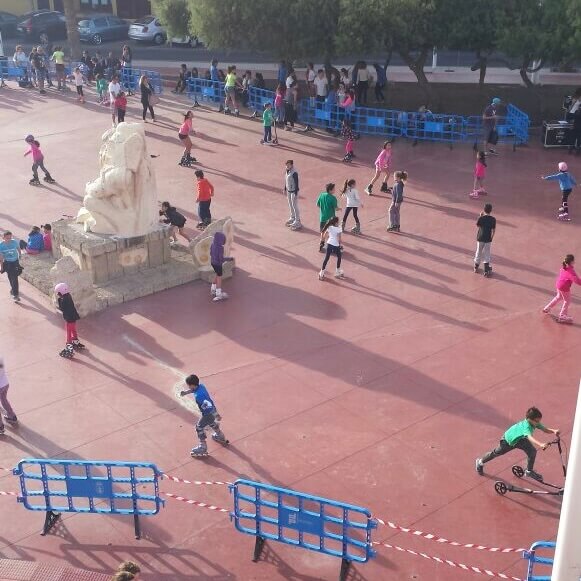 Niños patinando en una plaza, vistos desde un balcón, junto a una escultura de piedra y vallas azules.